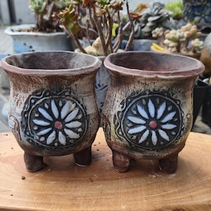 May include: Two small, brown, three-legged ceramic planters with a weathered, textured finish. Each planter features a prominent, circular relief design on the front, showcasing a white, multi-petaled flower motif with a small, central red dot.