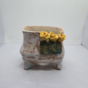 Op de afbeelding: Een kleine keramische plantenbak op pootjes met een verweerde witte en terracotta afwerking. De voorzijde is voorzien van een decoratief rechthoekig paneel met drie gebeeldhouwde gele rozen en groene bladeren aan de bovenrand.