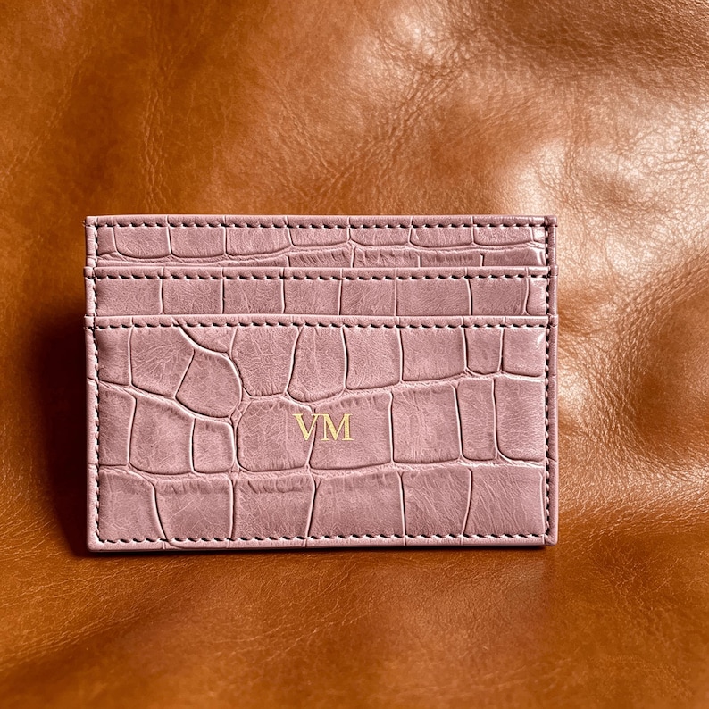 May include: A rectangular card holder in a dusty pink hue featuring a textured crocodile skin pattern. The front surface displays the gold-colored embossed initials VM centered on the lower portion, with visible stitching along the edges.