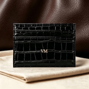 May include: A black rectangular card holder with a glossy crocodile-embossed texture. The front features the gold-colored letters VM centered in the middle. The item has multiple horizontal slots for organizing cards and a slim, flat profile.