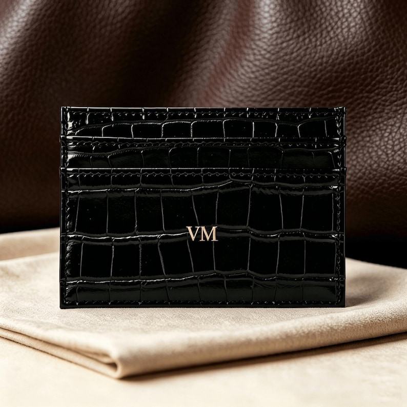 May include: A black rectangular card holder with a glossy crocodile-embossed texture. The front features the gold-colored letters VM centered in the middle. The item has multiple horizontal slots for organizing cards and a slim, flat profile.