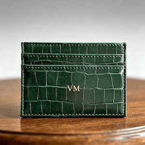 May include: A dark green card holder featuring a textured crocodile skin pattern and gold embossed initials VM on the front. The rectangular accessory includes multiple horizontal slots for organizing cards and has visible stitching along the edges.