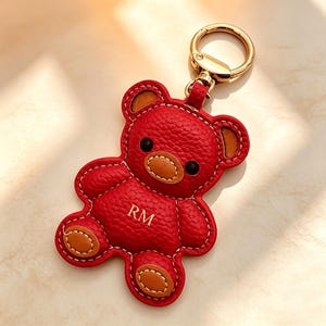 May include: Red leather teddy bear keyring with gold-coloured hardware. The bear has brown accents on the ears, nose, and feet, with the initials "RM" stitched on the front. The keyring is designed with a gold-coloured clasp.