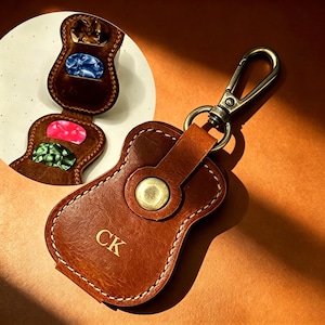 Può includere: Un portaplettri in pelle marrone a forma di chitarra con chiusura a scatto in ottone e le lettere CK impresse sul davanti. L'accessorio è dotato di un moschettone in metallo e contiene quattro plettri colorati blu, rosa e verdi.