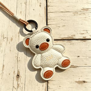 May include: A cream-colored leather teddy bear keychain with brown accents on the ears, nose, and paws. The keychain has a black ring and a brown cord. The bear is stitched with white thread.