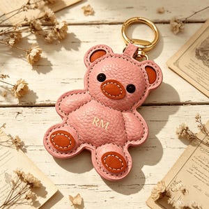 May include: Pink leather teddy bear keychain with brown accents and gold-tone hardware. The bear has the initials "RM" on its chest. The keychain is on a light wood surface with dried flowers and vintage paper.