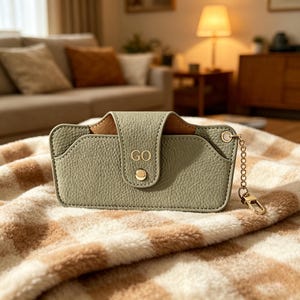 May include: A sage green textured case with a flap closure secured by a gold-toned snap button. The front features the letters GO embossed in gold. A gold-toned metal chain with a lobster clasp is attached to the right side of the rectangular case.
