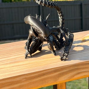 Può includere: Scultura nera stampata in 3D di una Regina Alien, con un esoscheletro dettagliato e una lunga coda segmentata. La scultura è posizionata su una superficie di legno, mostrando il suo design intricato e la sua postura minacciosa.