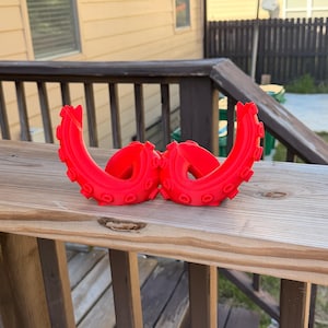 Puede incluir: Dos objetos rojos brillantes con forma de tentáculo, posiblemente decorativos o funcionales, se exhiben en una barandilla de madera. Los objetos tienen un diseño curvo con detalles circulares, que se asemejan a tentáculos de pulpo. El fondo muestra una casa y una valla.