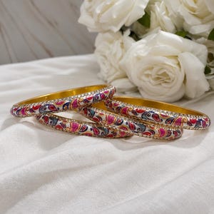 May include: Three gold-toned metal bangles featuring a white enamel surface decorated with a colourful paisley pattern in shades of pink, blue, and gold, accented by a border of small, clear, reflective stones along the top and bottom edges.