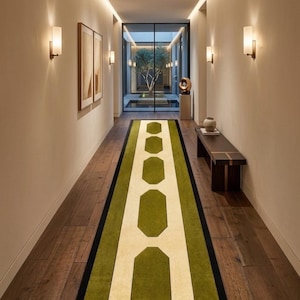 May include: A long rectangular runner rug featuring a central cream-colored stripe with a series of green octagonal shapes, bordered by solid green panels and a thin black outer edge, laid out on a dark wood floor in a hallway.
