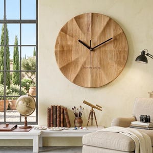 Può includere: Un orologio da parete rotondo in legno marrone chiaro con una superficie strutturata e sfaccettata. L'orologio presenta lancette nere per ore e minuti, una lancetta dei secondi nera e il testo "Enjoy the slow life" in piccole lettere nere.