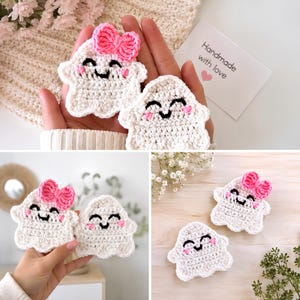 May include: Two small, white crocheted ghost figures with black embroidered smiling faces and pink stitched cheeks. One ghost features a bright pink crocheted bow on its head. The figures are crafted with a textured yarn finish and have a rounded, scalloped base.