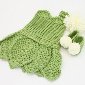 Può includere: Un completo per neonati all'uncinetto verde chiaro fatto a mano, che include un vestito con dettagli a forma di foglia, una fascia abbinata e un paio di stivaletti con pompon bianchi. Il completo è per un bambino.