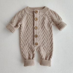 May include: A beige long-sleeve knitted romper featuring a textured diamond pattern, a ribbed crew neckline, and ribbed cuffs on the sleeves and legs. The front placket is secured with five round, light brown wooden buttons down the center.