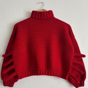 May include: A vibrant red, handmade crochet sweater with a turtleneck and unique sleeve detailing. The sleeves feature vertical, open slits, adding a distinctive design element to this cozy garment. The sweater is displayed on a wooden hanger.