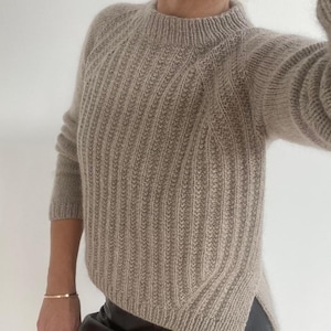 Può includere: Un maglione beige a maniche lunghe caratterizzato da un motivo a maglia a coste verticali, un collo alto rotondo e un orlo asimmetrico con spacchi laterali. Il capo è realizzato in un filato morbido e soffice con maniche raglan e vestibilità.