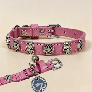 Op de afbeelding: Een roze leren halsband voor huisdieren met zilverkleurige metalen schedel- en kruisemblemen. De halsband is voorzien van een zilveren gesp, een klein belletje en een ronde metalen naamplaatje met de naam Mia en een roze pootafdruk.