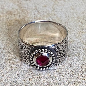 May include: A wide silver band ring featuring a textured, hammered surface. The center of the ring is adorned with a single, faceted, deep red gemstone set within a circular frame decorated with small, raised silver beads around its perimeter.