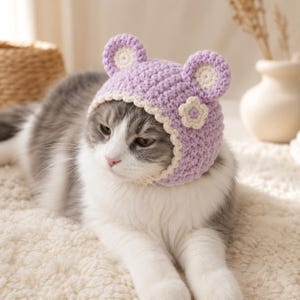 May include: A light purple crochet hat featuring two rounded ears with cream-colored centers and a small cream-colored flower accent on the side. The hat has a scalloped cream-colored border that frames the face, crafted from textured yarn.