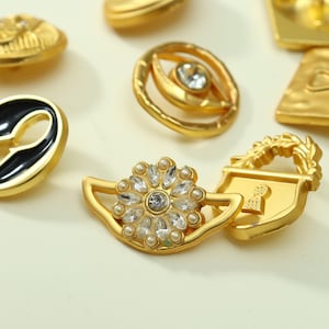 Pode incluir: Uma coleção de botões de metal em tom dourado com vários designs, incluindo uma peça circular com um cristal transparente central, um botão em forma de cadeado com detalhe de fechadura e um botão floral adornado com cristais transparentes e pérolas.