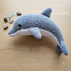 May include: A plush dolphin toy crafted from soft gray and white yarn with a textured, velvety finish. The dolphin features a prominent dorsal fin, side flippers, and a white underbelly. It has a single dark, glossy eye visible on the side.