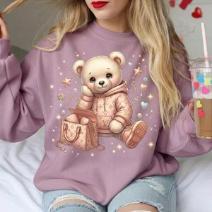 May include: A mauve crewneck sweatshirt featuring a graphic of a light brown teddy bear wearing a tan hoodie and matching sneakers. The bear sits next to a tan handbag, surrounded by small stars, hearts, and pearls on the front of the garment.