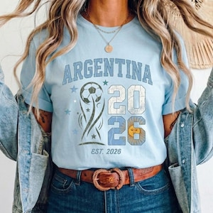 May include: A light blue t-shirt featuring the text ARGENTINA in a distressed font, a soccer ball graphic, the number 2026 with a sun icon inside the zero, and the text EST. 2026. The design includes small star accents in shades of blue.