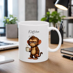 Könnte beinhalten: Ein weißer Keramikbecher mit einer Cartoon-Illustration eines braunen Affen mit müdem Gesichtsausdruck, der eine kleine Tasse dampfenden Kaffee hält. Über dem Affen steht der Text (K)äffchen in einer schwarzen, kursiven Handschrift-Schriftart.