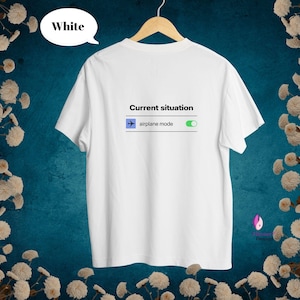 Può includere: Una t-shirt bianca a maniche corte appesa a una gruccia di legno. La maglietta presenta il testo nero Current situation sopra la grafica di un interruttore per la modalità aereo con un'icona di aereo blu e un indicatore attivo verde.