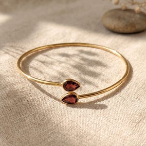 Puede incluir: Una pulsera rígida abierta de tono dorado con dos piedras preciosas de color rojo oscuro en forma de lágrima en los extremos. La fina banda de metal pulido se curva en forma circular, con las dos piedras enfrentadas y un pequeño espacio.