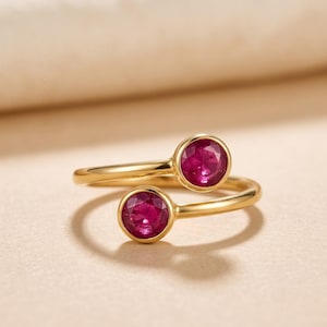 Peut inclure: Une bague ouverte en or ornée de deux pierres précieuses rondes serties clos, d'une couleur rouge rosée vive. L'anneau en or poli s'enroule autour du doigt, chaque extrémité se terminant par un sertissage circulaire accueillant une pierre facettée.