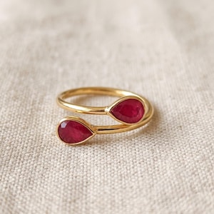 Può includere: Un anello aperto in oro con due pietre preziose rosse sfaccettate a forma di goccia, fissate a ciascuna estremità della fascia. La montatura in metallo dorato lucido si curva per sostenere le due vivaci pietre rosse.