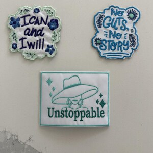 May include: Three embroidered patches on a white background. The top left patch is scalloped with blue flowers and the text "I CAN and I will". The top right patch is blue and white with the text "No GUTS No STORY". The bottom rectangular patch features a teal outline of a person in a wide-brimmed hat and the word "Unstoppable".