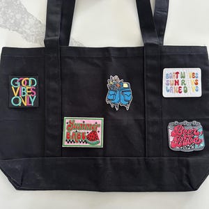 May include: A black canvas tote bag featuring five embroidered patches. The patches display the text "GOOD VIBES ONLY", "BOAT WAVES SUN RAYS LAKE DAYS", and "Summer BABE" with a watermelon, a pair of blue denim pockets, and a pink beverage can.