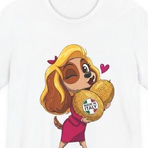 Può includere: Una t-shirt bianca con un'illustrazione a cartone animato di un cane marrone e beige con capelli biondi, che indossa un completo rosa, fa l'occhiolino e tiene in mano due meloni. Un melone ha un adesivo circolare con scritto MADE IN ITALY e la bandiera italiana.