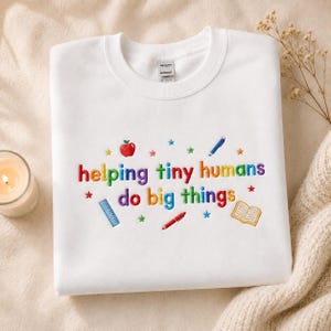 May include: A white crewneck sweatshirt featuring colorful embroidered text that reads "helping tiny humans do big things." The design includes small embroidered icons of a red apple, a blue ruler, a red pen, a blue pencil, an open book, and stars.