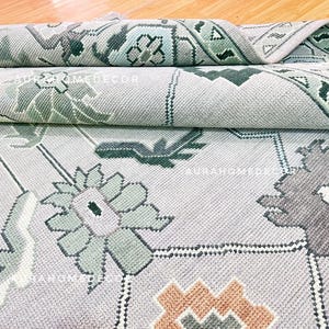 Puede incluir: Una alfombra de color gris claro con un patrón geométrico y floral repetitivo en tonos verde salvia, gris carbón oscuro y naranja apagado. La alfombra tiene un tejido texturizado e incluye el texto AURAHOMEDECOR impreso en la superficie.