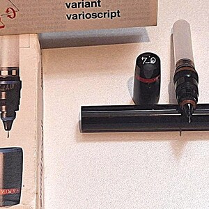 Puede incluir: Un bolígrafo técnico de dibujo con cuerpo blanco, agarre negro y punta metálica fina. El bolígrafo viene con una tapa negra marcada con el número 0.4, una tapa roja pequeña y un estuche cilíndrico negro. El embalaje muestra el texto variant varioscript.