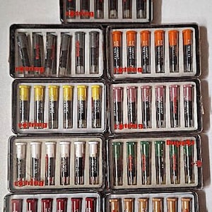 May include: A collection of nine rectangular plastic cases containing Rotring technical pen ink cartridges. Each case holds six individual cartridges with color-coded caps, including brown, black, orange, yellow, pink, white, green, red, and tan.