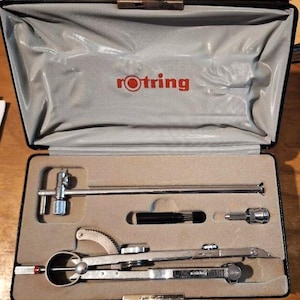 Könnte beinhalten: Ein offener schwarzer Koffer mit grauem Innenraum, der Zeichenwerkzeuge enthält. Die Werkzeuge sind silber und schwarz, darunter ein Zirkel, ein technischer Stift und andere Zeicheninstrumente. Das rote "rotring"-Logo ist auf dem Koffer sichtbar.