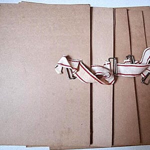 Può includere: Un set di cartelle portadocumenti rettangolari in cartone color marrone chiaro con finiture in metallo e lacci in tessuto bianco sporco con sottili strisce rosse. Le cartelle sono impilate e legate con i lacci in tessuto per chiuderle.