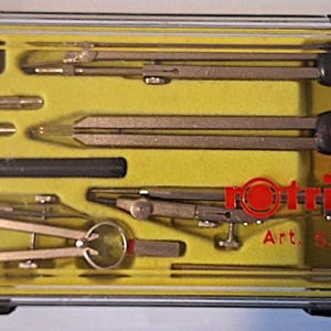 Könnte beinhalten: Ein rechteckiger, durchsichtiger Kunststoffkasten mit gelbem Innenraum, der ein Set aus silbernen Metallzirkeln und technischen Zeichengeräten enthält. Das rote rotring-Logo und der Text Art. 530216 sind auf dem Deckel aufgedruckt.
