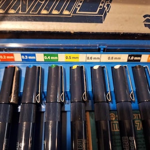 May include: A set of black fine-liner pens with silver clips, arranged in a blue case. The pens have colored tips and are labeled with sizes ranging from 0.1 mm to 1.2 mm. The brand name "STA" is visible.