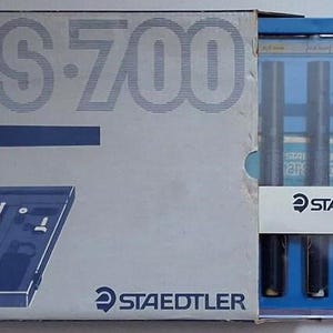 Peut inclure: Un ensemble de stylos techniques Staedtler Mars 700 dans un étui en carton gris et bleu. L'emballage porte le texte mars 700 et Staedtler. À l'intérieur du boîtier en plastique transparent se trouvent quatre stylos noirs et des accessoires.