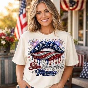 Puede incluir: Una camiseta de color crema con un gráfico de labios con el patrón de la bandera estadounidense en rojo, blanco y azul, decorado con estrellas y fuegos artificiales brillantes. Debajo, el texto Star Spangled Mama en letra cursiva brillante.