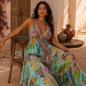 Puede incluir: Una mujer con un vestido largo con cuello halter y un estampado floral vibrante en tonos turquesa, rosa y amarillo. El vestido tiene un escote pronunciado y un panel central decorativo. El escenario es al aire libre, con muebles de madera rústicos.