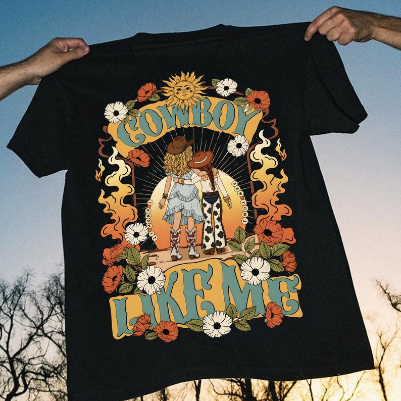 Pu&ograve; includere: Una t-shirt nera con una stampa grafica di due figure con cappelli e stivali da cowboy. Il design include un sole, fiori e fiamme in arancione, giallo e bianco, con il testo "COWBOY LIKE ME" scritto in grandi lettere di colore blu.