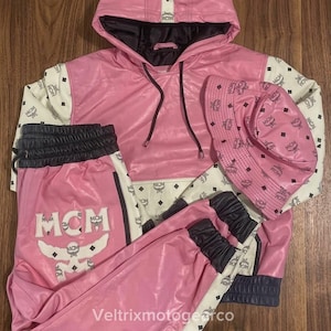Puede incluir: Un conjunto de sudadera con capucha, pantalones de chándal y sombrero de pescador en rosa y blanco. Las prendas presentan un patrón repetido del logotipo MCM, cordones negros y cinturas y puños elásticos negros. El pantalón lleva un logo MCM grande.