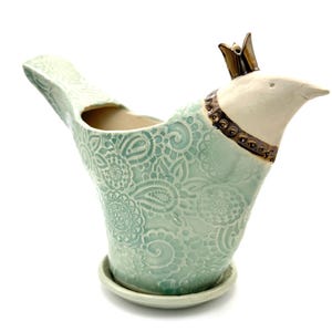 May include: A ceramic bird-shaped planter in a pale mint green color with an embossed lace-like floral pattern. The bird has a white head, a metallic bronze-colored crown, and a matching bronze collar. It sits on a small, round matching saucer.